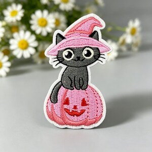 Puede incluir: Un parche bordado que muestra un gato gris oscuro con un sombrero de bruja rosa claro, sentado sobre una calabaza de Halloween rosa claro con una cara tallada en rojo. El diseño tiene un borde blanco y un estilo de dibujos animados.