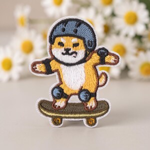 Puede incluir: Un parche bordado que muestra a un perro Shiba Inu de color canela y blanco con un casco azul, coderas negras y rodilleras negras mientras monta en un monopatín de color verde oliva oscuro. El diseño tiene un borde blanco y costuras.