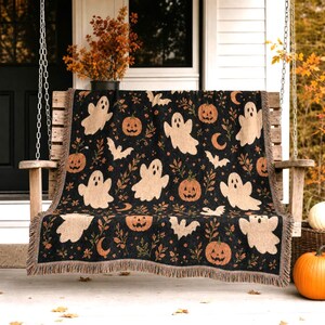 May include: A black woven throw blanket featuring a repeating pattern of beige ghosts, orange jack-o'-lanterns, small bats, crescent moons, and autumn foliage. The rectangular blanket has a fringed edge and a textured, tapestry-style construction.