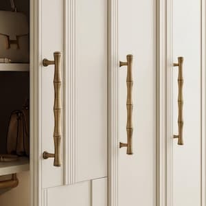 May include: A tall, off-white wardrobe featuring vertical paneling and long, metallic brass-toned handles with a segmented bamboo-like texture. The open section reveals shelves holding handbags and a clothing rod with wooden hangers inside.