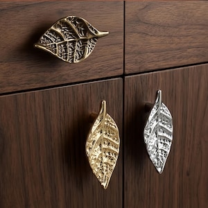 Puede incluir: Tres pomos de metal para armarios con forma de hoja, fijados en paneles de madera oscura. Un pomo tiene un acabado de latón antiguo, otro un acabado dorado pulido y el tercero un acabado plateado pulido, con texturas de vetas detalladas.