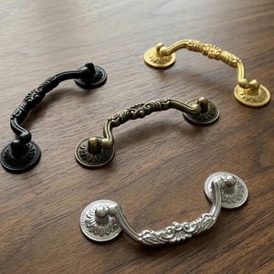 May include: Four metal drawer pulls in black, antique brass, bronze, and silver finishes. Each handle features a curved, ornate design with intricate floral detailing along the grip and circular mounting plates with a petal-like pattern.