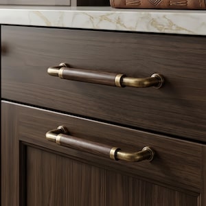 May include: Two horizontal drawer pulls featuring a dark brown wood grain center section flanked by brushed antique brass metal ends. The hardware is mounted onto dark wood cabinet drawers with a visible horizontal grain pattern and clean lines.