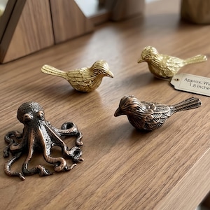 May include: Three metal figurines including two birds and one octopus. One bird is gold-toned, one bird is copper-toned, and the octopus is copper-toned. A small white tag attached to the gold bird displays the text Approx. Width 1.8 inches.