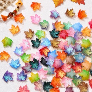 Puede incluir: Una colección de pequeños dijes con forma de hoja de arce en una variedad de colores translúcidos y brillantes, incluyendo naranja, verde, rosa, azul, morado y dorado. Cada pieza presenta una textura de venas detallada y un pequeño orificio.