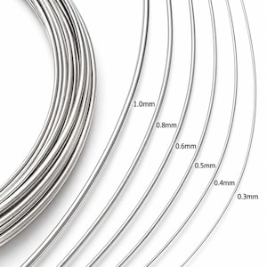 Könnte beinhalten: Eine Spule aus silberfarbenem Metalldraht in verschiedenen Stärken, beschriftet mit den Maßen 1.0mm, 0.8mm, 0.6mm, 0.5mm, 0.4mm und 0.3mm. Der Draht ist glatt, reflektierend und kreisförmig angeordnet, um die verschiedenen Stärken zu zeigen.