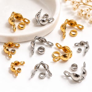 May include: A collection of gold and silver-toned metal jewelry findings, specifically clamshell bead tips and small crimp beads. These components feature a hinged, rounded design with loops for attachment, arranged in various pairs across the surface.