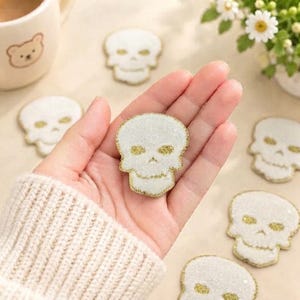 May include: A collection of white skull-shaped patches with gold glitter accents on the eye sockets and edges. Each patch features a textured, sparkling surface. The skull patches are arranged on a light-colored surface, with one held in the palm.
