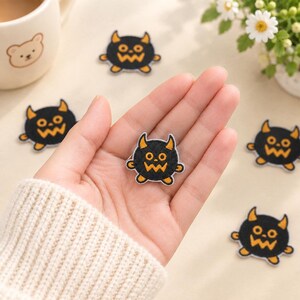 May include: A small, circular embroidered patch featuring a black monster character with orange horns, orange eyes, and an orange jagged mouth. The patch has a white border and is designed with a flat, stitched texture for application to fabric items.