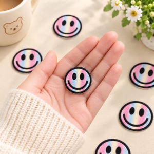 May include: A circular embroidered patch featuring a gradient rainbow pattern with a black smiley face design. The patch has a black stitched border and is shown in a palm, with several identical patches scattered on a light surface nearby.