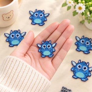 Puede incluir: Varios parches bordados termoadhesivos pequeños con forma de monstruos azules con alas, ojos blancos grandes y vientres azul claro. Cada parche presenta un borde azul oscuro, una superficie texturizada y un aspecto de dibujos animados.