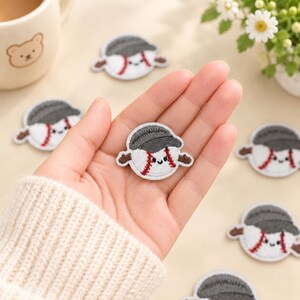May include: A circular embroidered patch featuring a white baseball with red stitching and a smiling face, wearing a dark grey baseball cap. The patch has small brown arms extending from the sides, creating a character design with a friendly expression.