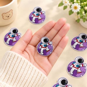 May include: A circular embroidered patch featuring an astronaut in a white suit with red and blue accents, walking on a purple cratered moon surface. The patch is circular with a white border, showcasing detailed stitching of the space explorer.