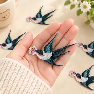 May include: Multiple embroidered swallow patches featuring dark blue wings, white underbellies, and red throat accents. Each bird is depicted in flight, carrying a small, light pink flower in its beak. The patches are detailed with intricate thread work.