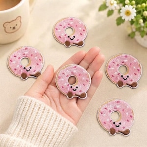 May include: Five circular embroidered patches shaped like pink donuts with brown sprinkles and smiling faces. Each patch features a central hole, light pink icing, and small brown details representing arms or feet, arranged on a light surface.