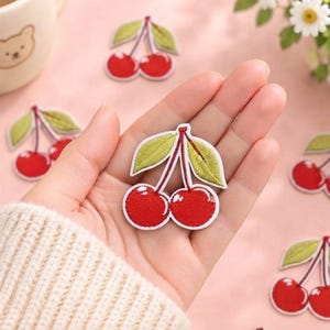 May include: An embroidered cherry patch featuring two bright red cherries with white highlights, connected by thin stems to two textured light green leaves. The patch has a white border and is held in a palm against a soft pink background.