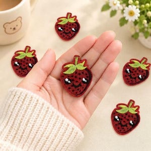 May include: Five small, red strawberry-shaped embroidered patches. Each patch features a green stem, a dotted texture, and a smiling face with two white eyes and a black mouth. One patch is held in the palm, while four others are scattered nearby.