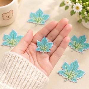 May include: Several small, flat, die-cut paper maple leaf shapes in shades of teal, turquoise, and light green. Each leaf features a detailed, darker green vein pattern printed on the surface. The leaves are arranged in a scattered, loose formation.