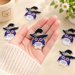 Puede incluir: Un parche bordado de un búho morado con un sombrero de bruja negro, una hebilla amarilla y un detalle de telaraña blanca. El búho tiene ojos grandes blancos, un pico amarillo y patas blancas. Varios parches de búho idénticos están dispuestos.
