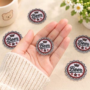 Op de afbeelding: Een ronde geborduurde patch in de vorm van een kroonkurk met een wit midden, een rode binnenring en een donkergrijze geschulpte rand. De patch bevat het woord Beer in zwarte schrijfletters, een klein bruin kroontje en drie rode sterren.