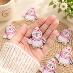 May include: A small embroidered patch featuring a penguin wearing a pink knit hat and a matching pink scarf. The penguin has a white belly, grey wings, and orange feet. The patch is designed with detailed stitching to create a textured appearance.