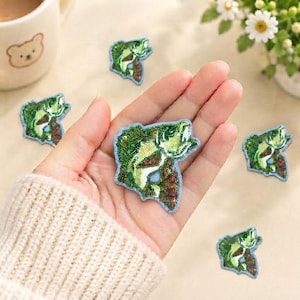 May include: Several small, embroidered iron-on patches shaped like green and white largemouth bass fish. Each patch features detailed stitching of a fish in a jumping pose with a light-colored belly and dark green scales, finished with a light blue border.