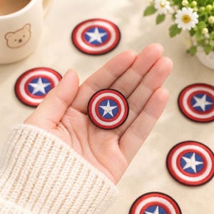 Op de afbeelding: Een ronde geborduurde patch met een rood, wit en blauw schildontwerp met een centrale witte ster. De patch heeft een zwarte rand en wordt in een hand gehouden, met verschillende identieke patches verspreid over een lichtgekleurd oppervlak.