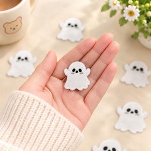 Puede incluir: Un pequeño parche bordado con forma de fantasma blanco con ojos y boca cosidos en negro. El fantasma tiene una superficie texturizada y un borde inferior festoneado. Varios parches de fantasmas idénticos están dispersos alrededor del central.