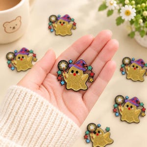 May include: An embroidered patch featuring a golden ghost wearing a purple witch hat, holding a sparkler, and surrounded by small colorful stars. The ghost has two black eyes and a red mouth, with detailed stitching throughout the design.