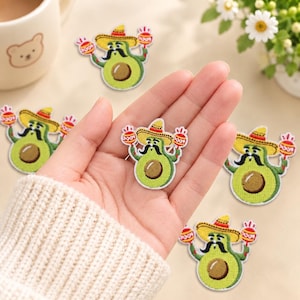 May include: An embroidered iron-on patch featuring a green avocado character wearing a yellow sombrero and a black moustache, holding red and white maracas in each hand. The avocado has a brown pit in the centre and a white border around the edge.