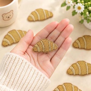 May include: A collection of small, golden-brown croissant-shaped embroidered patches. Each patch features detailed stitching that mimics the layered texture of a pastry. The patches are scattered across a light-colored surface.