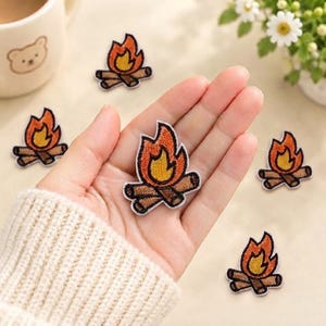May include: Multiple embroidered campfire patches featuring orange and yellow flames atop brown logs. Each patch has a white border and is designed for fabric application. The design depicts a classic bonfire with detailed stitching.