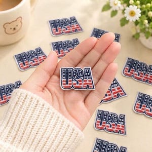 以下が含まれることがあります： 太字でUSAと書かれた刺繍ワッペン。各ワッペンは赤、白、青の配色で、Uの文字には星柄、SとAの文字には赤と白の横縞模様が施されています。これらは衣類や布製品に縫い付けたりアイロンで接着したりするための装飾的なアップリケです。