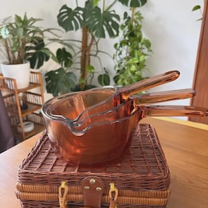 May include: A set of three nested amber-colored glass saucepans with integrated handles and pouring spouts. The translucent, warm-toned glass vessels are stacked together, showcasing a smooth, rounded design with a consistent deep orange hue.