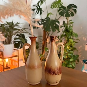Peut inclure: Deux vases en céramique de style burette avec anses, présentant une base beige clair et un vernis dégradé brun plus foncé autour du col et de la section médiane. Un vase contient une tige de plante séchée plumeuse sortant de son ouverture.