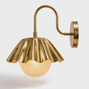 May include: A wall-mounted lamp featuring a polished brass finish with a curved arm and a circular wall plate. The light fixture includes a wide, ruffled, metallic shade that surrounds a spherical, frosted white glass globe light bulb.