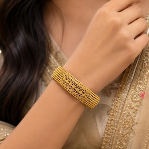 May include: A wide gold bangle bracelet featuring an intricate filigree pattern of repeating heart-shaped motifs, bordered by rows of small, textured gold beads along the top and bottom edges, creating a detailed and structured metallic design.