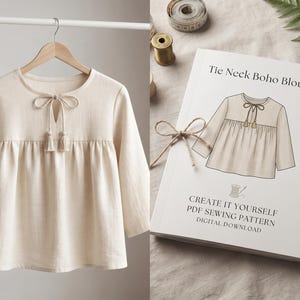 Op de afbeelding: Een crèmekleurige blouse met lange mouwen, een ronde halslijn, een split aan de voorzijde en een striksluiting met twee kwastjes. Daarnaast ligt een wit boekje met de tekst Tie Neck Boho Blouse, Create It Yourself PDF Sewing Pattern.