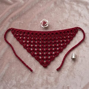 Pode incluir: Uma bandana triangular de crochê em vermelho escuro com um padrão de losangos abertos e laços longos nas extremidades. Um pequeno ornamento de rosa branca e rosa está posicionado acima do centro e um dedal de metal prateado à direita.