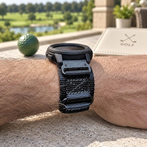 May include: A black digital smartwatch featuring a rugged, textured nylon strap with a metal buckle attachment. The watch case is circular and matte black, secured to the wrist with a heavy-duty woven band that includes reinforced stitching details.