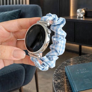 May include: A smartwatch with a round, black digital display and a polished silver metal casing. The watch band is a gathered, scrunchie-style fabric strap featuring a light blue and white gingham checkered pattern, attached to the watch face.