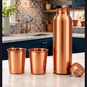 Puede incluir: Un juego de tres objetos de color cobre que consta de dos vasos cilíndricos y una botella de agua alta de cuello estrecho con su tapa de rosca a juego colocada al lado sobre una superficie de mármol blanco.