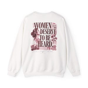 Può includere: Una felpa a maniche lunghe color bianco sporco con una grafica bordeaux sul retro. Il design include il testo "WOMEN DESERVE TO BE HEARD", l'illustrazione di una persona, elementi floreali, un edificio e il testo "EVERY VOICE EVERY STORY EVERY WOMAN."