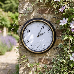 Puede incluir: Un reloj de pared redondo con marco negro y dorado, que presenta una esfera blanca con números arábigos negros, manecillas negras ornamentadas y un termómetro integrado en la parte inferior. El reloj muestra claramente los números del uno al doce.