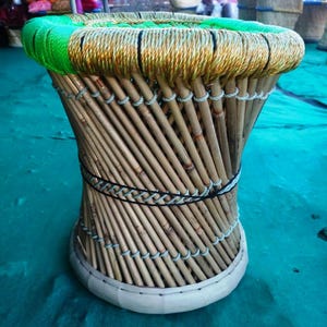 May include: A round stool constructed from vertical bamboo poles arranged in a tapered hourglass shape. The top edge is wrapped in bright green and metallic gold cord, while a black cord secures the center. The base is finished with a white band.