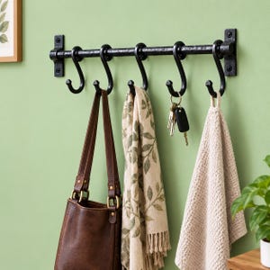 May include: A black metal wall-mounted rack featuring a horizontal bar with five removable S-shaped hooks. The rack holds a brown leather bag, a beige scarf with a green leaf pattern, a set of metal keys, and a textured beige towel.