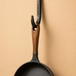 May include: A black cast iron skillet with a wooden handle hanging from a dark, textured metal hook. The skillet features the number 26 embossed on the neck near the handle. The hook is secured to the wall with a single visible black screw.