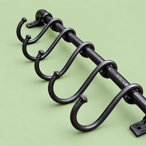 May include: A dark gray metal wall-mounted rack featuring a horizontal bar with five removable S-shaped hooks. The rack has two flat mounting plates at each end, each with a single circular hole for attachment to a wall surface.