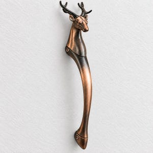Puede incluir: Un tirador de puerta vertical hecho de metal con un acabado en cobre y bronce oscuro. La parte superior del tirador presenta una cabeza de ciervo esculpida con astas, mientras que la sección inferior se curva hacia abajo hasta una base redondeada con tornillos visibles.