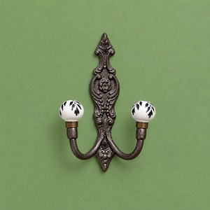 May include: A double wall hook featuring a dark bronze-toned metal backplate with ornate scrollwork and floral detailing. Two curved arms extend outward, each topped with a white ceramic knob decorated with a black leaf pattern.